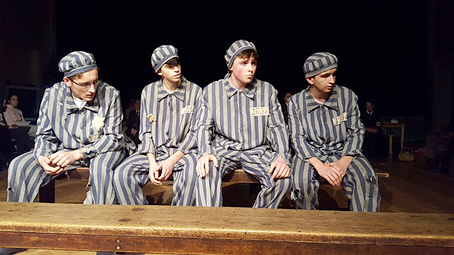 A scene showing Andreas Frohlich and 3 fellow prisoners, seated, wearing the Mauthausen concentration camp's striking prison uniform (with prominent vertical stripes, and matching cap)